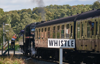 Steam tain whistle This landscape photograph, taken during a bright autumn morning in 2018, features a steam train as the main subject, with an emphasis on the prominent "WHISTLE" sign in the foreground. The scene is set in the rural surroundings of the North Yorkshire Moors in the United Kingdom, capturing the classic charm of rail transport as the steam train moves along the track. The image shows the traditional carriages reflecting the greenery and early autumn foliage, while railway staff interact near the engine and a passenger leans from a carriage window. The photograph highlights the continuing significance of historic train travel in this rural setting, encapsulating the heritage of steam train journeys in the North Yorkshire Moors.